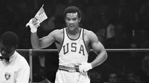 Bettmann via Getty Images Foreman wearing a top with the word 'USA' on the front, waving a US flag