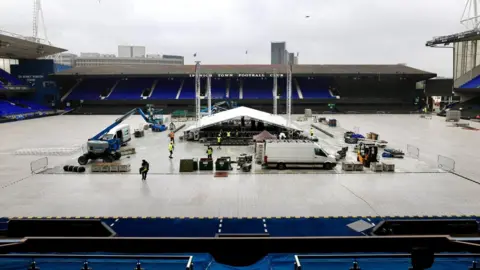 Queensberry Promotions A large metal structure with a white roof, which all looks like a big tent, is in place in the middle of the pitch. It is surrounded by the football stadium's stands. Workers in hi-vis jackets are working around it and various vehicles are parked nearby. The pitch is covered in white protective boards.