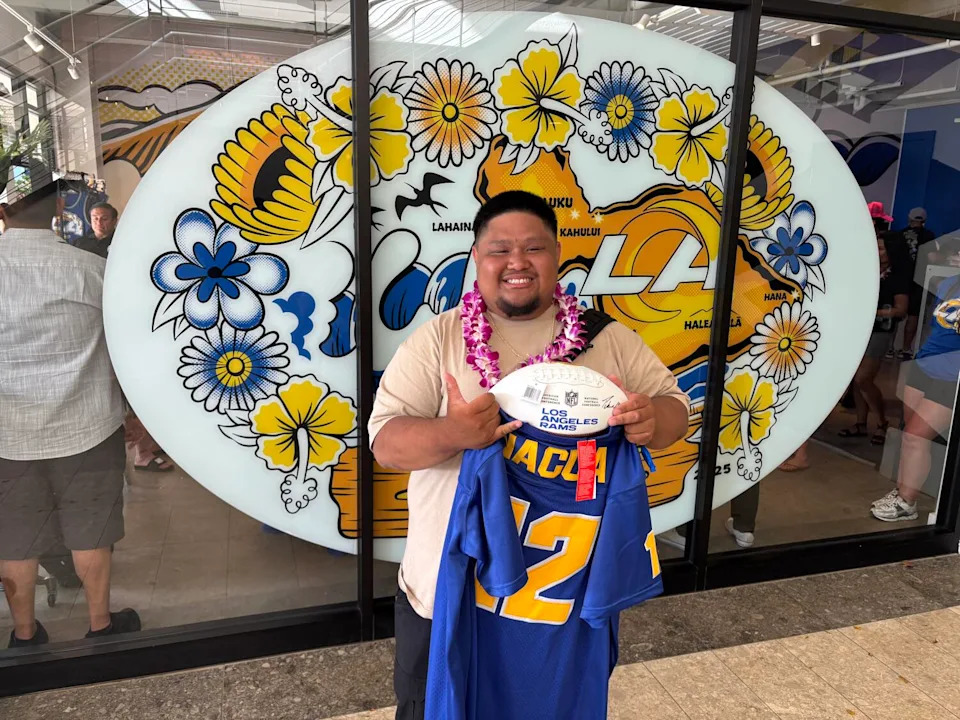 Joshua Cabjuan, wearing a lei, does the 'hang loose' hand sign while smiling with a copy of Nacua's high school jersey