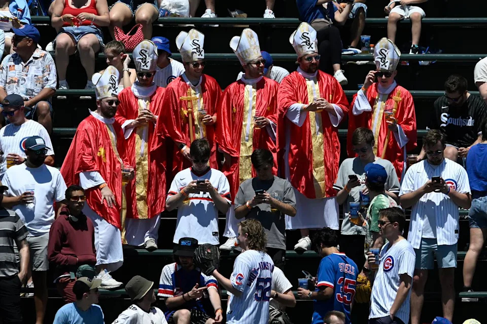 Six fans wearing red and gold robes and white mitres with White Sox logos in the stands among other baseball fans