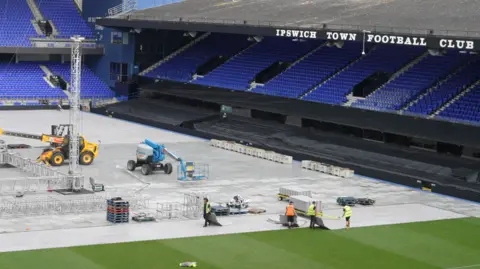 Jamie Niblock/BBC Workers wearing hi-vis jackets walking on the pitch at Portman Road. The pitch is mostly covered in protective plastic boards, but part of the playing surface remains exposed. There is a yellow heavy machinery vehicle putting large metal structures in place where the boxing ring will be. A stand with blue seats is on the right of the picture.