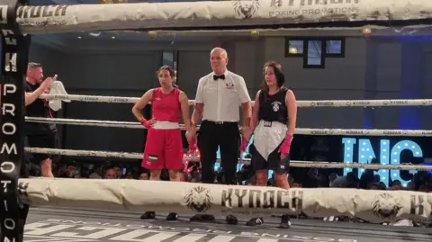 Bravehearts Amateur Boxing Club Two female boxers - in red kit on the left and black on the right - stand either side of a boxing referee in white shirt and black bow tie, in the boxing ring at the end of the bout.