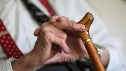 PA Media A close of an elderly man's hands. He is holding a walking stick and wearing a white shirt and a tie. 
