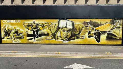 NATHAN MURDOCH STREET ARTS HIRE PETERBOROUGH A close up of part of a mural, painted in yellow, black and white, showing Walter Cornelius, face on and flat on his stomach with his hands beneath his shoulders. His head is raised and he is looking forward and in front of him on the ground is a pea.