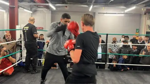 RICHARD KNIGHTS/BBC Khan, who is wearing a grey T-shirt and black trousers, is holding his hands up which have boxing gloves on. A person is standing in front of him with their back to the camera wearing red boxing gloves.