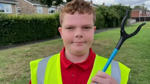 JOHN DEVINE/BBC A head and shoulders shot of a boy with short gingery hair, wearing a hi-vis yellow waistcoat over a red polo-shirt. He is holding a blue-handled litter picker up to his shoulder. Behind him is grass, a hedge and some just glimpsed houses. 