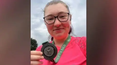 ASHER COOPER Asher Cooper who is taking a selfie and looking directly at the camera and smiling. She has a medal round her neck and is holding this up and showing the camera. She is wearing a hot pink running top and is wearing glasses.