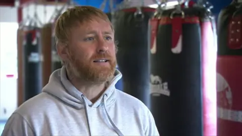 BBC Stefy Bull, who has strawberry blonde hair and a ginger bear. Wearing a grey hoodie, he is stood in front of a boxing bag.