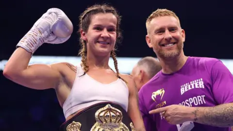 Getty Images A woman in white vest with a boxing glove raised smiles for the camera - it's Terri Harper, female world champion. Next to her Steffi Bull, in a purple T-shirt with sponsor logos on, points at Ms Harper.