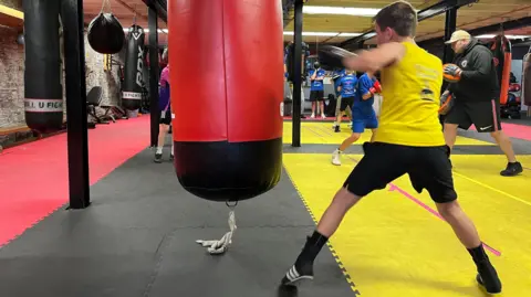 A boy in a yellow vest, black shorts and black boots punches a red punchbag.