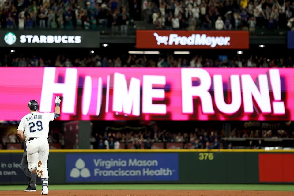 Cal Raleigh is seen from behind lifting his arm as he rounds the bases with 'Home Run' displayed on the scoreboard