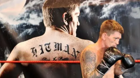 PA Media A wide shot of Ricky Hatton wearing black boxing gloves. In the background is an image of Hatton's back, with the tattoo Hitman written across it.