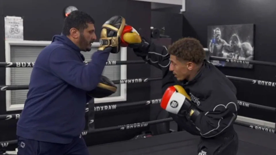 Saad is wearing a black tracksuit and red, white, yellow gloves. He is punching towards gold and black pads that his dad is holding. He is wearing a navy blue tracksuit. 