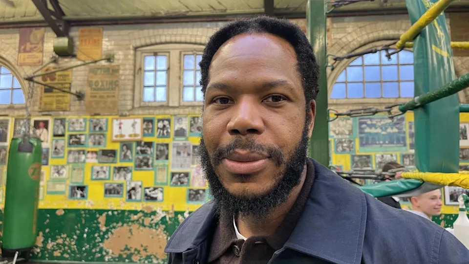 A man with dark hair and a beard stands in a boxing club. He is wearing a navy coat and a brown polo top with white T-shirt underneath. There are posters on the walls behind him and arched windows above. 