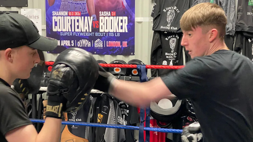 Two young men are in a boxing ring. One wears black cushioned hand pads, a black t-shirt and black cap. The other has short brown hair, a black t-shirt, black boxing gloves and is hitting the cushioned pads. In the background, there are many t-shirts and boxing posters pinned to the walls of the gym.