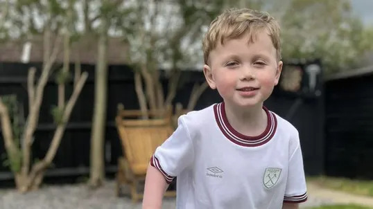 Jesse standing in his garden holding the crossbar of a small football goal. He is wearing a white West Ham kit and is smiling.