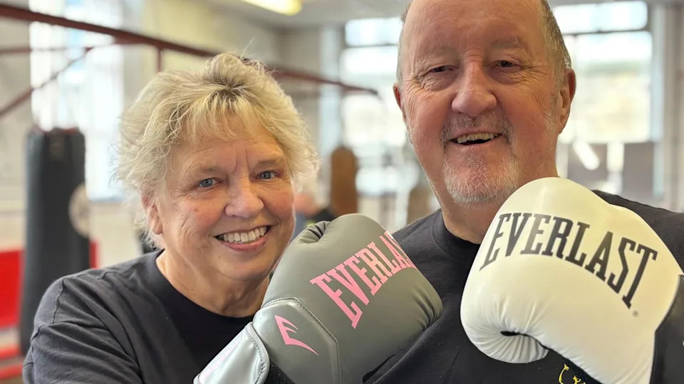 An elderly couple smile as they hold up their white boxing gloves to the camera.