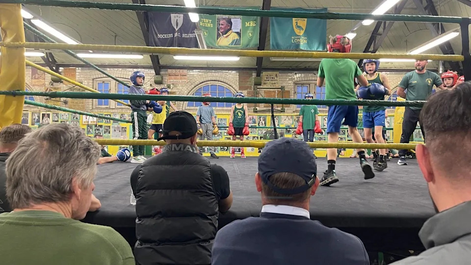 A group of primary school age children spar with each other in a boxing ring with green and gold ropes around it. Some men are sat in the club watching them from the sidelines. There are lots of pictures on the surrounding yellow walls. 