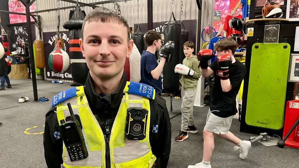A police officer stands in front of three young people in sportswear boxing inside a gym. The police officer has short black hair, blue lapels, a luminous vest, black uniform jacket, a walkie talkie and another gadget attached to his chest. Two of the three young people in the background are wearing boxing gloves and the other is wearing cushioned pads. There are punch bags hanging from a steel frame.