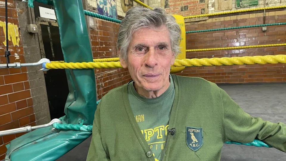 A man with thick grey hair is wearing a green cardigan with a logo that reads Repton A.B.C. He sits at the side of a boxing ring.
