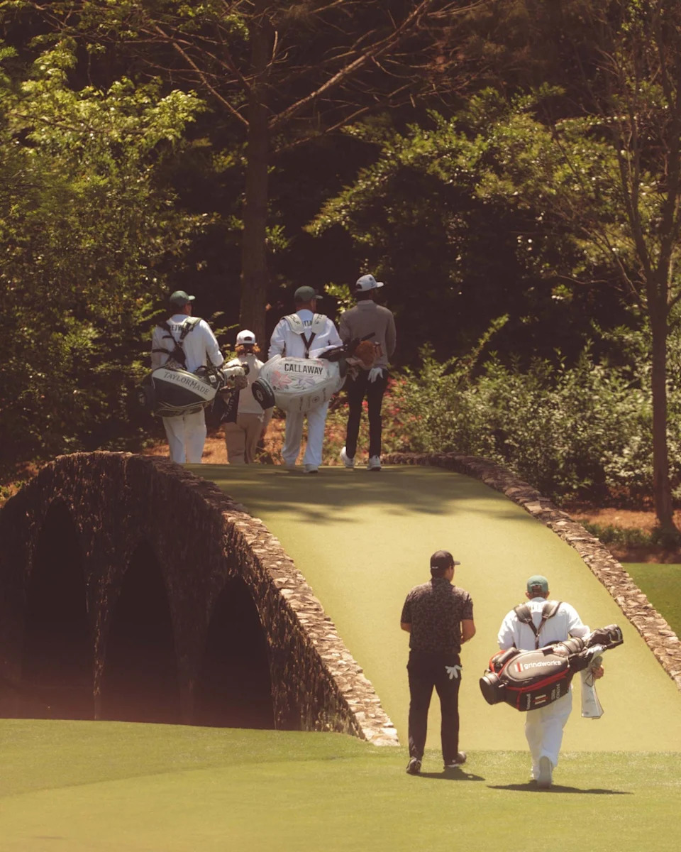 Golfers and caddies walk over a stone bridge on Augusta National’s lush golf course, surrounded by trees and greenery, with sunlight filtering through the foliage after having walked all 18.
