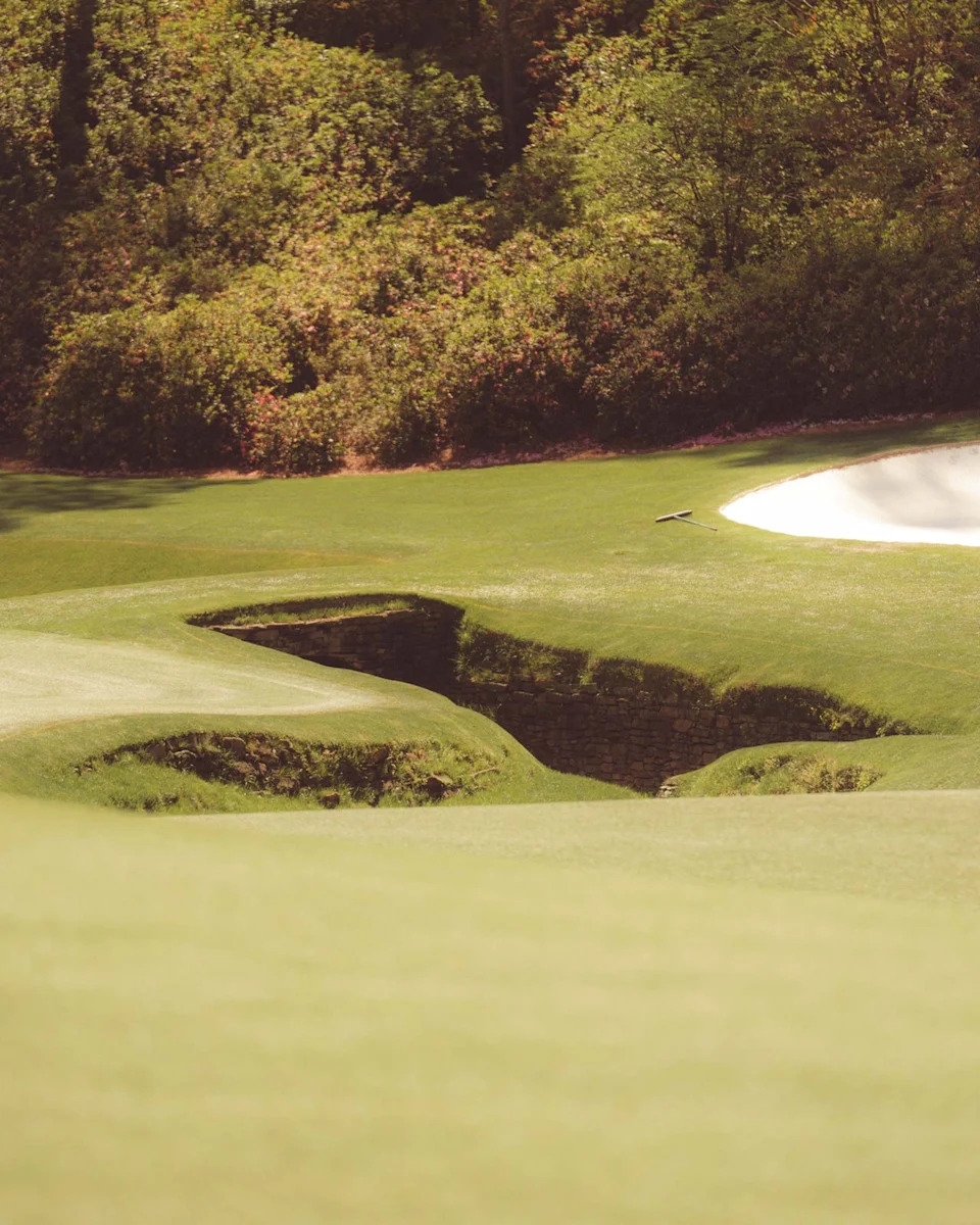 A golf course with a green fairway, a sand bunker on the right, and a small stone-lined creek running through the grass—reminiscent of Augusta National—surrounded by dense trees and shrubbery in the background.