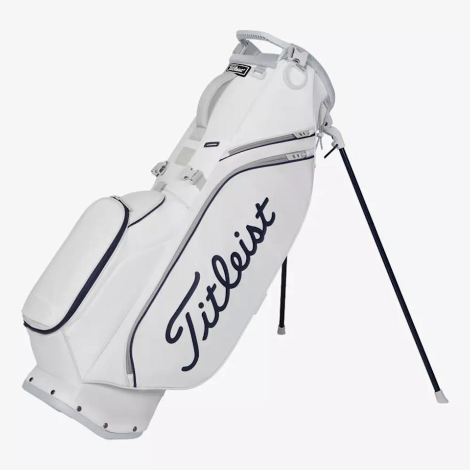 A white Titleist golf bag with navy blue accents and logo, featuring a stand and multiple zippered compartments, stands upright on its extended legs—a sleek contender among the best golf bags 2026.