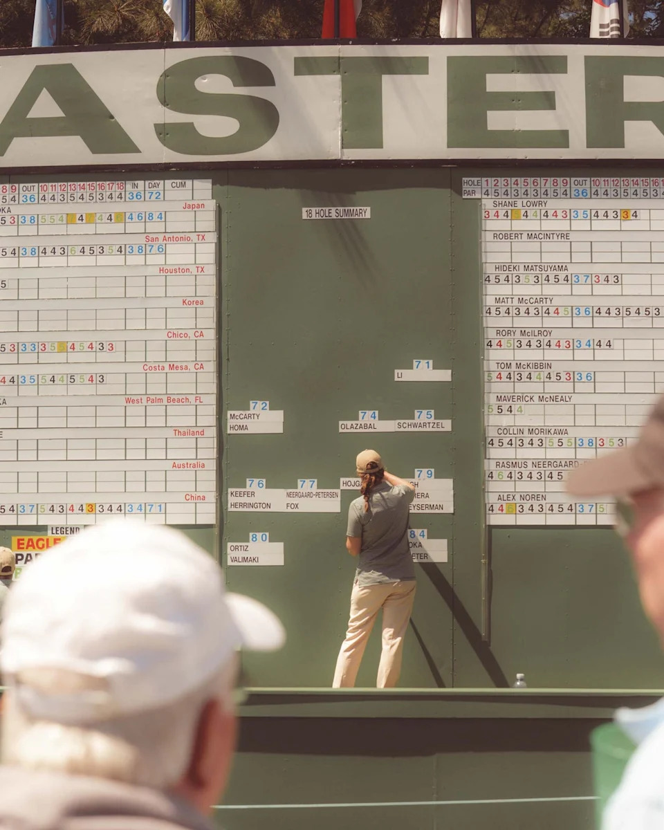 A person updates scores on a large, outdoor Masters golf tournament leaderboard at Augusta National while spectators watch from the foreground. Various player names and numbers are displayed on the board.