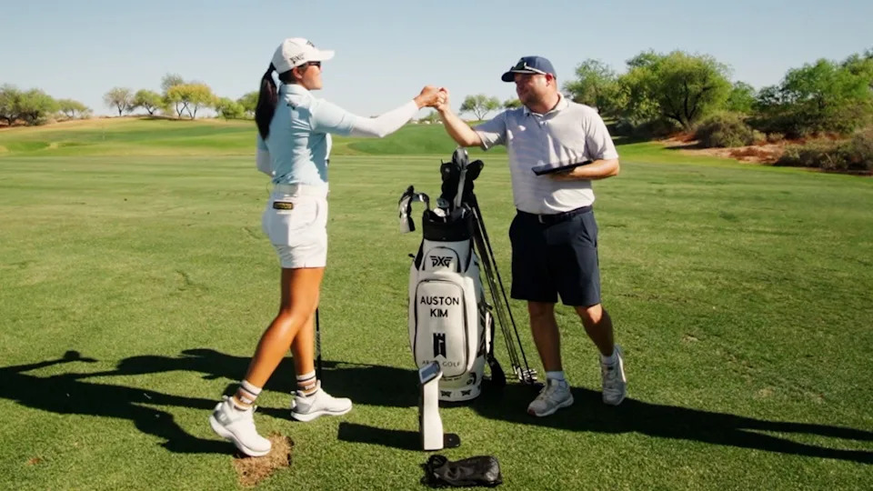 A female golfer and a male golfer fist bump next to a golf bag labeled “AUSTON KIM” on a sunny golf course. They stand on green grass surrounded by trees under a clear blue sky.