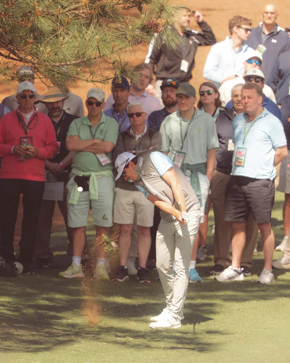 A golfer at Augusta National in mid-swing hits a shot from the grass near a tree, sending up a small spray of dirt, while spectators who walked all 18 holes stand closely behind, watching attentively.
