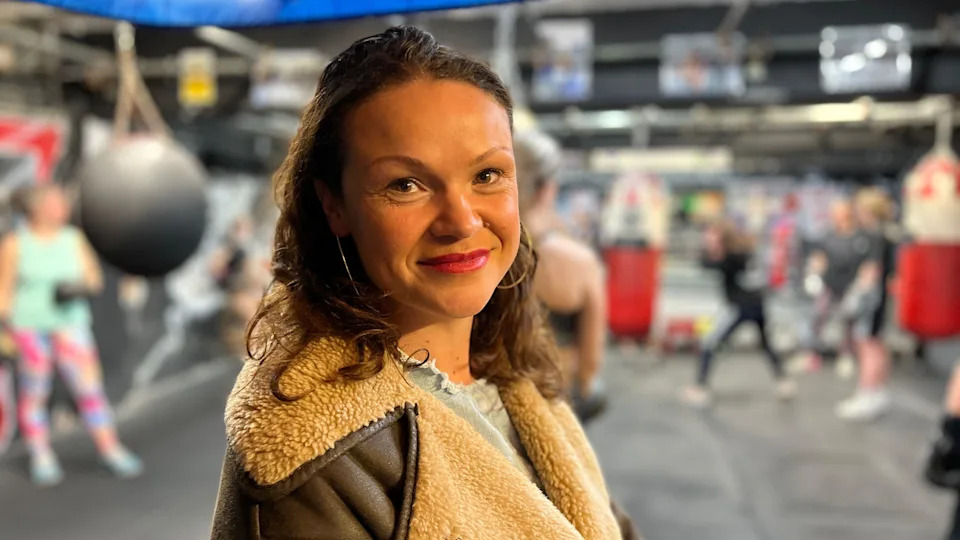 A women is turning to face the camera in a boxing gym. She is wearing a brown leather coat with a cream coloured sheepskin colour. She has shoulder length brown hair and is smiling. Women are training in the gym behind her. 