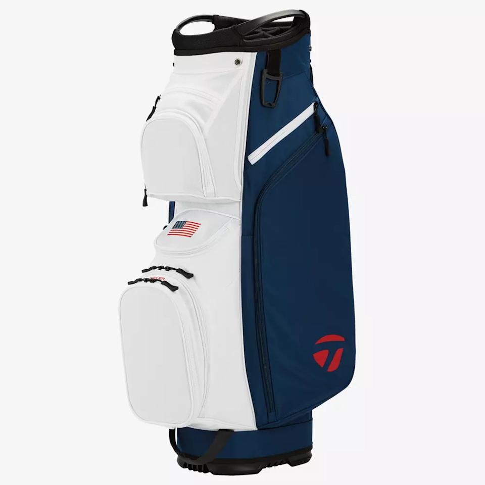 A golf bag with a white front, blue sides, several zippered pockets, an American flag patch, and a red logo near the base—an ideal choice for those seeking one of the best golf bags 2026—stands upright against a plain white background.