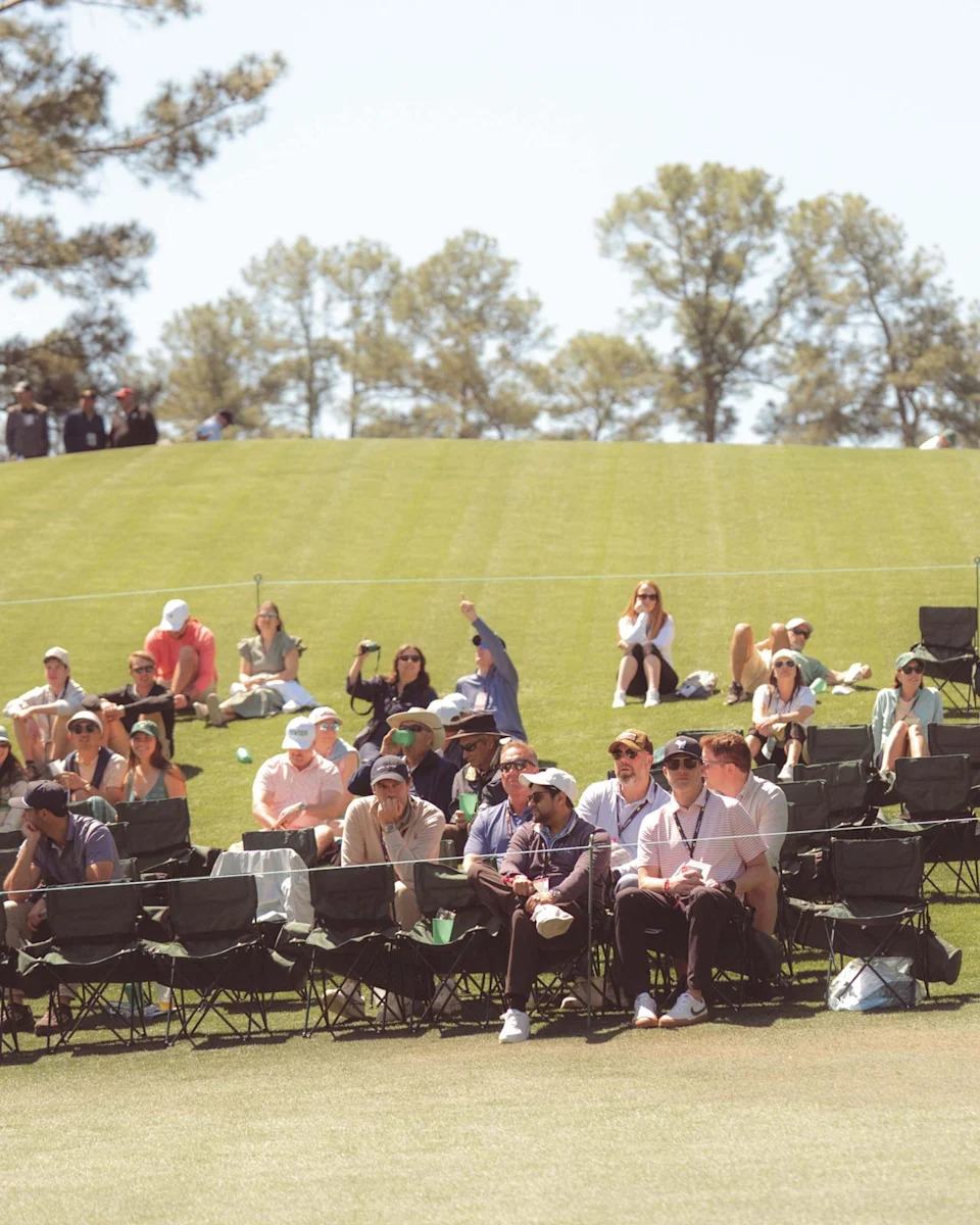 Spectators sit on foldable chairs and grass, watching a golf event on a sunny day. Trees and blue sky are visible in the background, evoking Augusta National vibes. Some wear hats and sunglasses, many relaxed—perhaps after having walked all 18 holes themselves.
