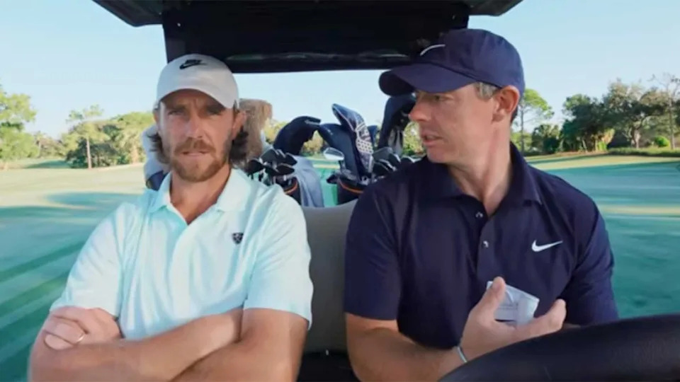 Two men, Rory and Tommy, sit side by side in a golf cart on a golf course, both wearing caps and collared shirts, golf clubs visible behind them. One man has his arms crossed while the other gestures in a classic Tale of the Tape moment.
