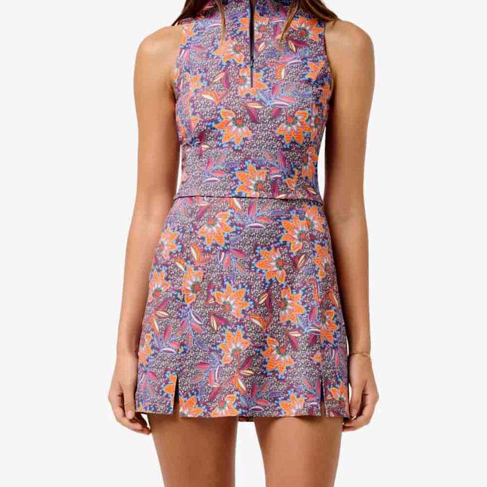 A woman wearing a sleeveless, high-neck, floral dress with a colorful pattern in purple, orange, and pink—a stylish choice for Mother’s Day. The dress features a fitted waist and short hemline with small front slits.