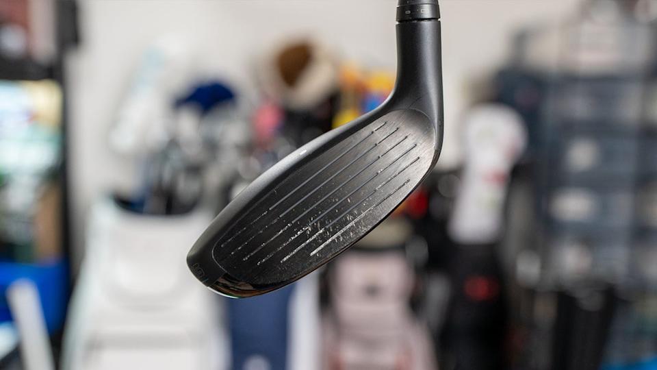 Close-up of a black golf club head with visible grooves and some wear marks, with a blurred background of various golf bags and equipment.