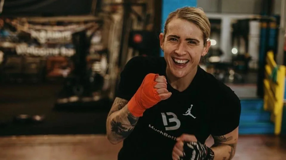 Jade, a woman with blonde hair, she wears a black t-shirt and has her hands strapped, she clenches her fist as if she is fighting and smiles brightly.