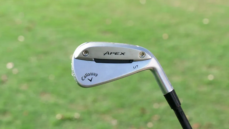 A close-up of a Callaway Apex UT golf iron with a silver head, photographed outdoors with a blurred green grass background.