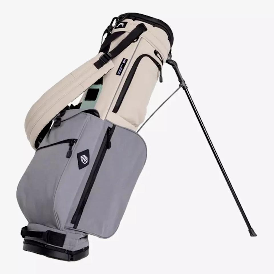 A beige and gray golf bag with a shoulder strap and two fold-out legs, designed to stand upright. Featuring multiple zippered pockets and a black logo, it's a stylish choice for those seeking the best golf bags 2026 has to offer.