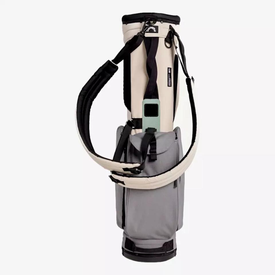 A minimalist golf bag in beige and gray with padded shoulder straps, front pocket, and a rectangular green tag by the top handle—this stylish contender stands upright on white and sets the standard for the best golf bags 2026.