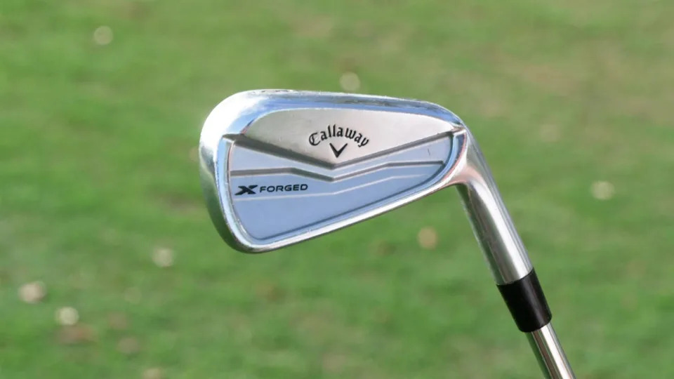 A close-up of a Callaway X Forged golf iron with a shiny silver finish, held against a blurred green grass background.