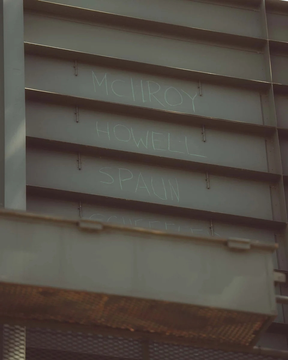 A metal wall with names MCILROY, HOWELL, and SPAUN written in white chalk across three panels—evoking the feel of Augusta National after you’ve walked all 18. A metal grate is partially visible at the bottom of the image.