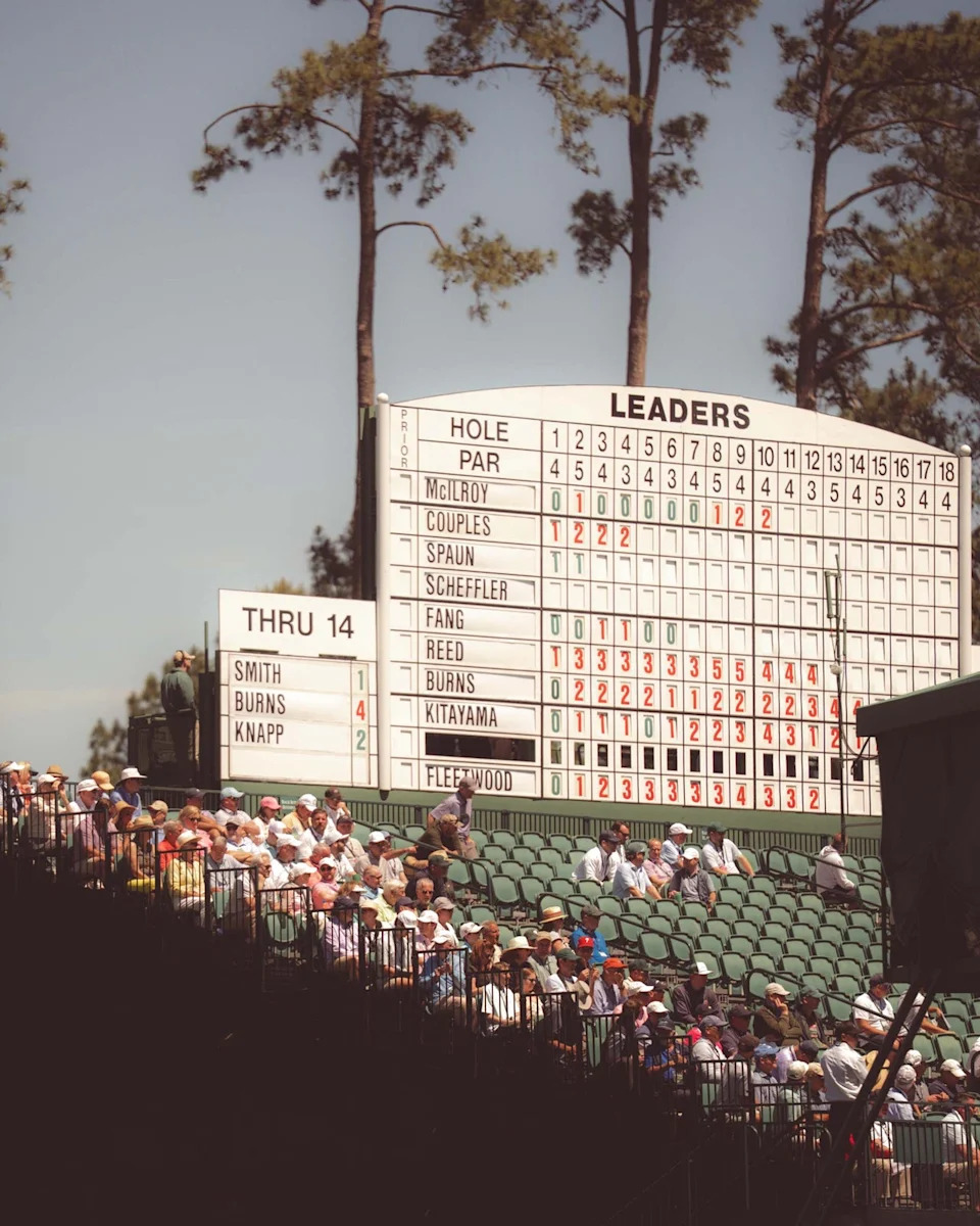 A crowd watches a golf tournament at Augusta National in front of a large leaderboard displaying players' names and scores, with tall pine trees in the background under a clear sky—many have walked all 18 holes to catch the action.
