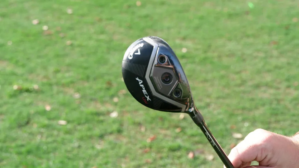 A person holds a Callaway Apex golf hybrid club outdoors over green grass.