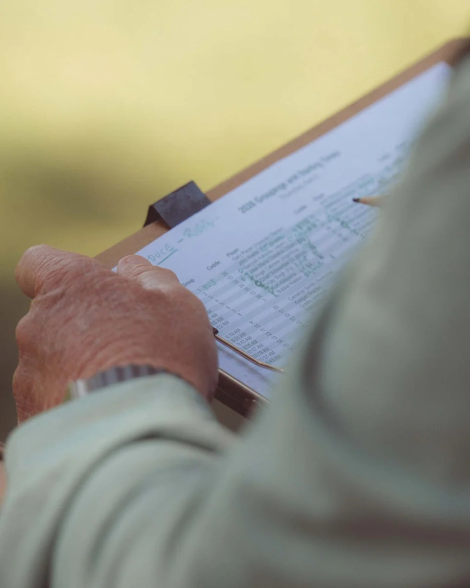 A person holding a clipboard with a printed form attached, appearing to review or fill out information—perhaps recording scores for a walk all 18 at Augusta National. The focus is on the hands and clipboard, with the background blurred.