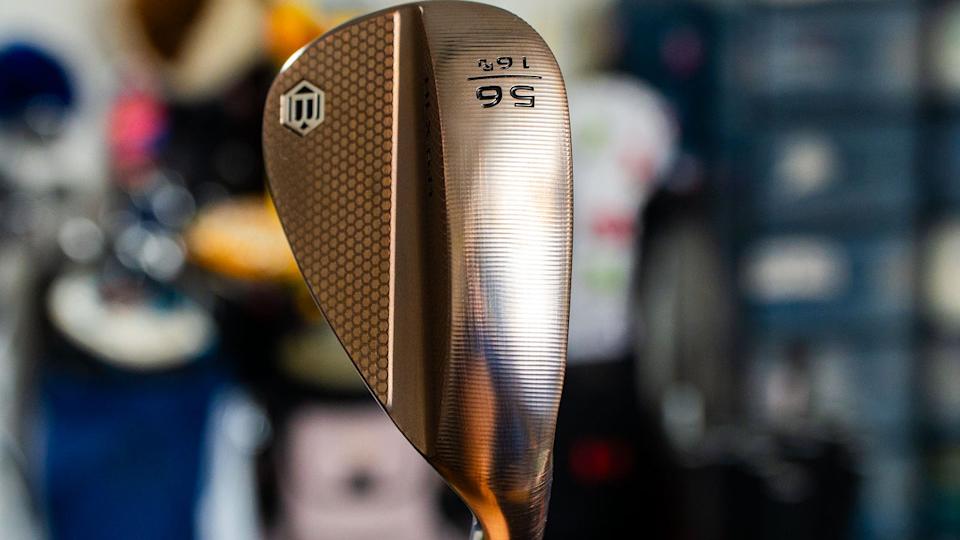 Close-up of a golf wedge with a bronze finish, showing the numbers 56 and 16 engraved on the club head, along with a logo on the textured surface. The background is blurred.