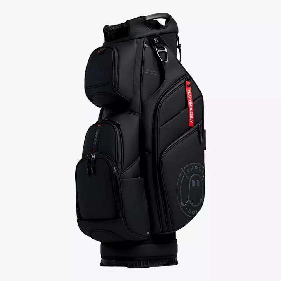 A black golf bag with multiple zippered compartments, a handle on top, a red tag reading FRAGMENT, and a subtle ghost logo on the lower side—an eye-catching contender for the best golf bags 2026. The bag stands upright against a plain white background.
