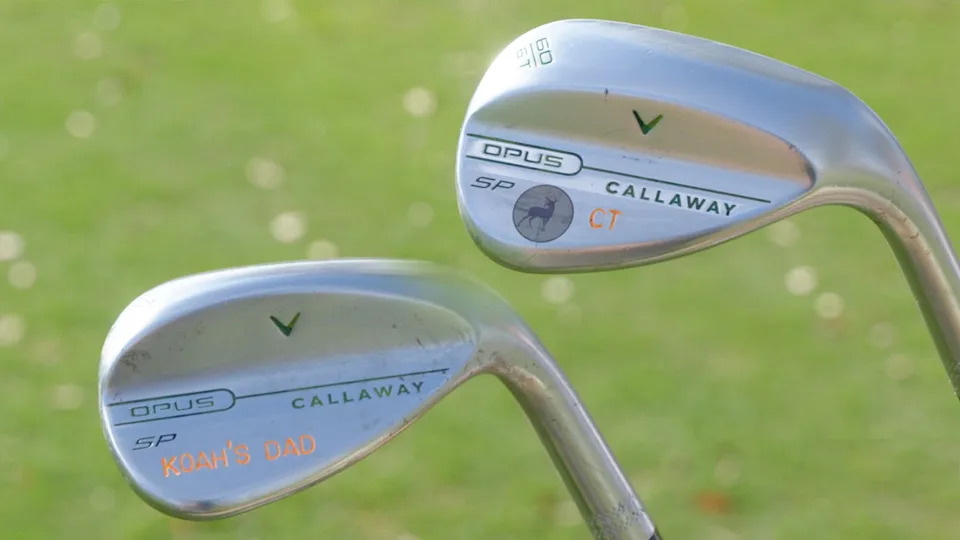 Two silver Callaway golf clubs with custom engravings on their heads rest upright on green grass. One club is labeled KOAHS DAD in orange lettering. Both clubs show the Opus and Callaway branding.