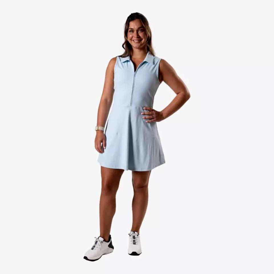 A woman stands smiling with one hand on her hip, wearing stylish apparel—a light blue sleeveless dress and white sneakers—against a plain white background, perfect for a Mother's Day look.