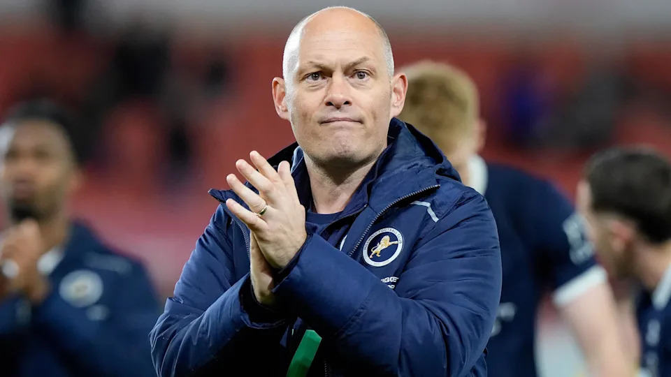 Alex Neil, wearing a navy blue Millwall jacket, claps his hands to applaud the fans after a win against Stoke